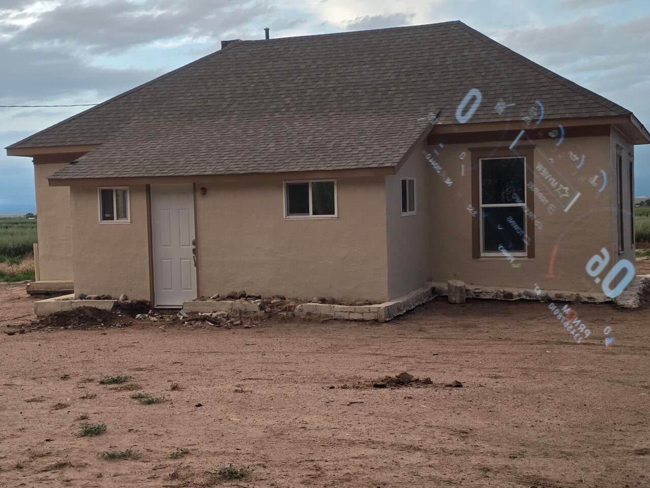 Single-story beige house with a dark roof on a bare dirt lot under a cloudy sky