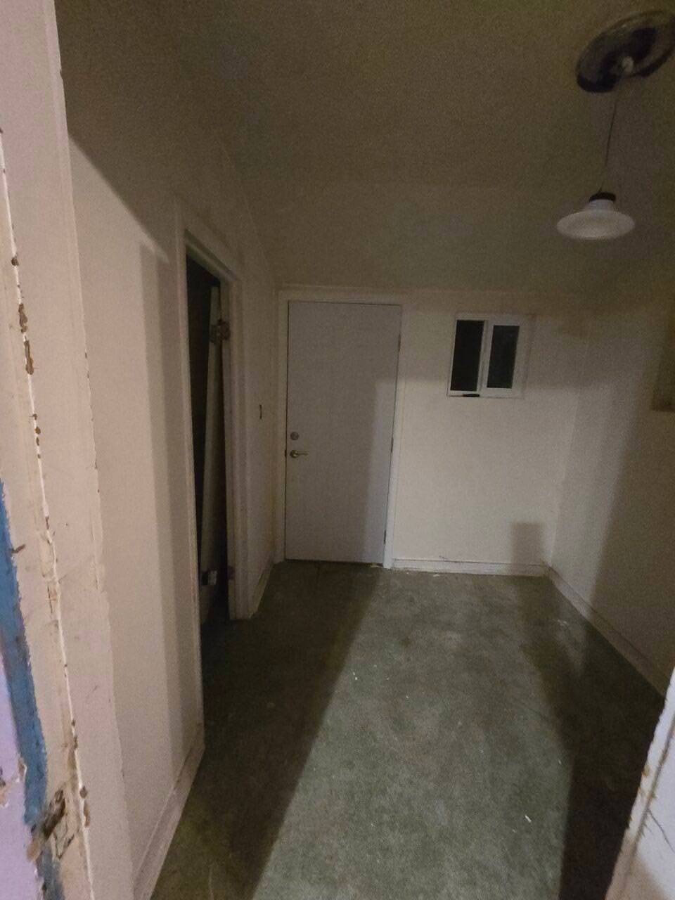 Empty narrow room with white walls, gray floor, closed door, and small window.