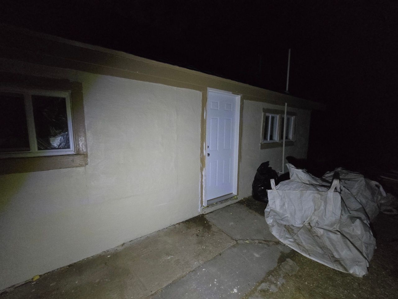 Nighttime view of a house exterior with a white door, windows, and covered items on the patio. Nighttime view of a house exterior with a white door, windows, and covered items on the patio.