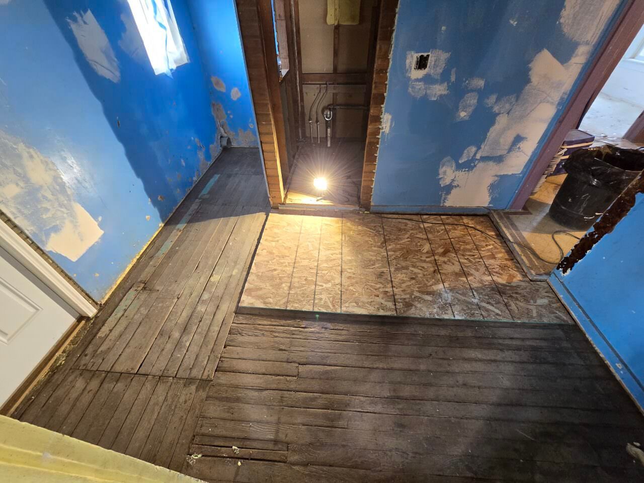 Dim unfinished room with blue walls, worn wooden floorboards, and a bright light at the far doorway