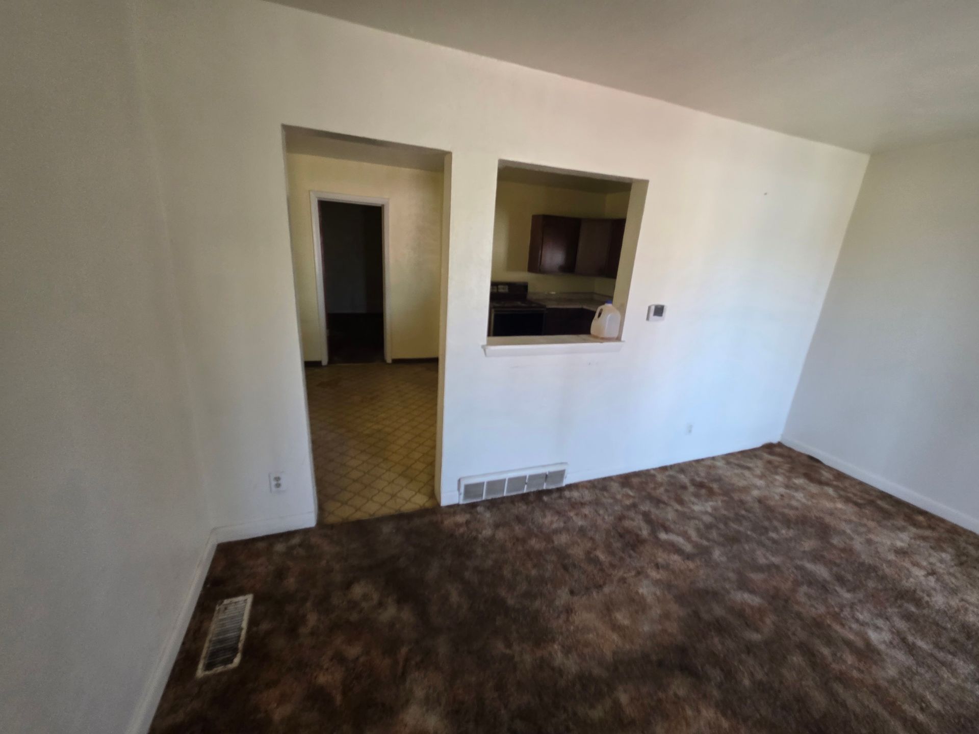 Empty unfurnished room with brown carpet, white walls, and a doorway opening to a kitchen pass-through. Empty unfurnished room with brown carpet, white walls, and a doorway opening to a kitchen pass-through.