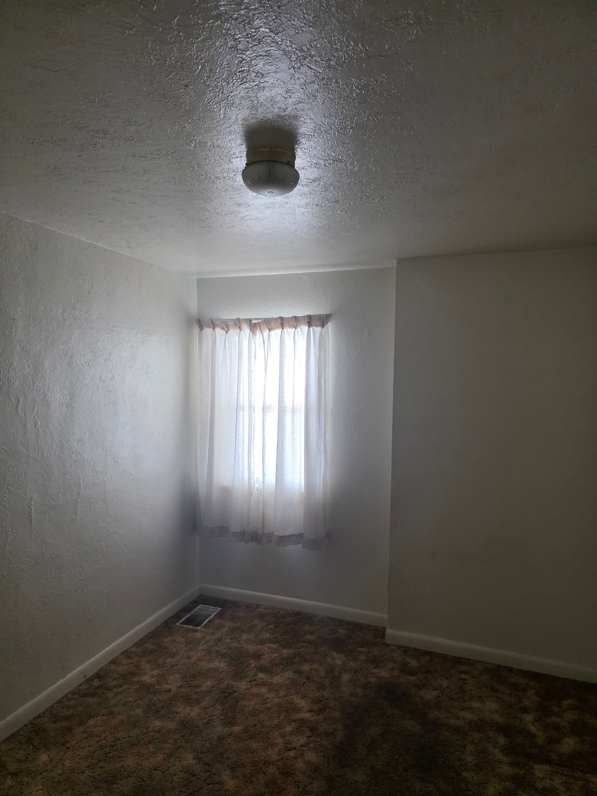 Empty room with textured white walls, a window with curtains, and a ceiling light fixture. Empty room with textured white walls, a window with curtains, and a ceiling light fixture.