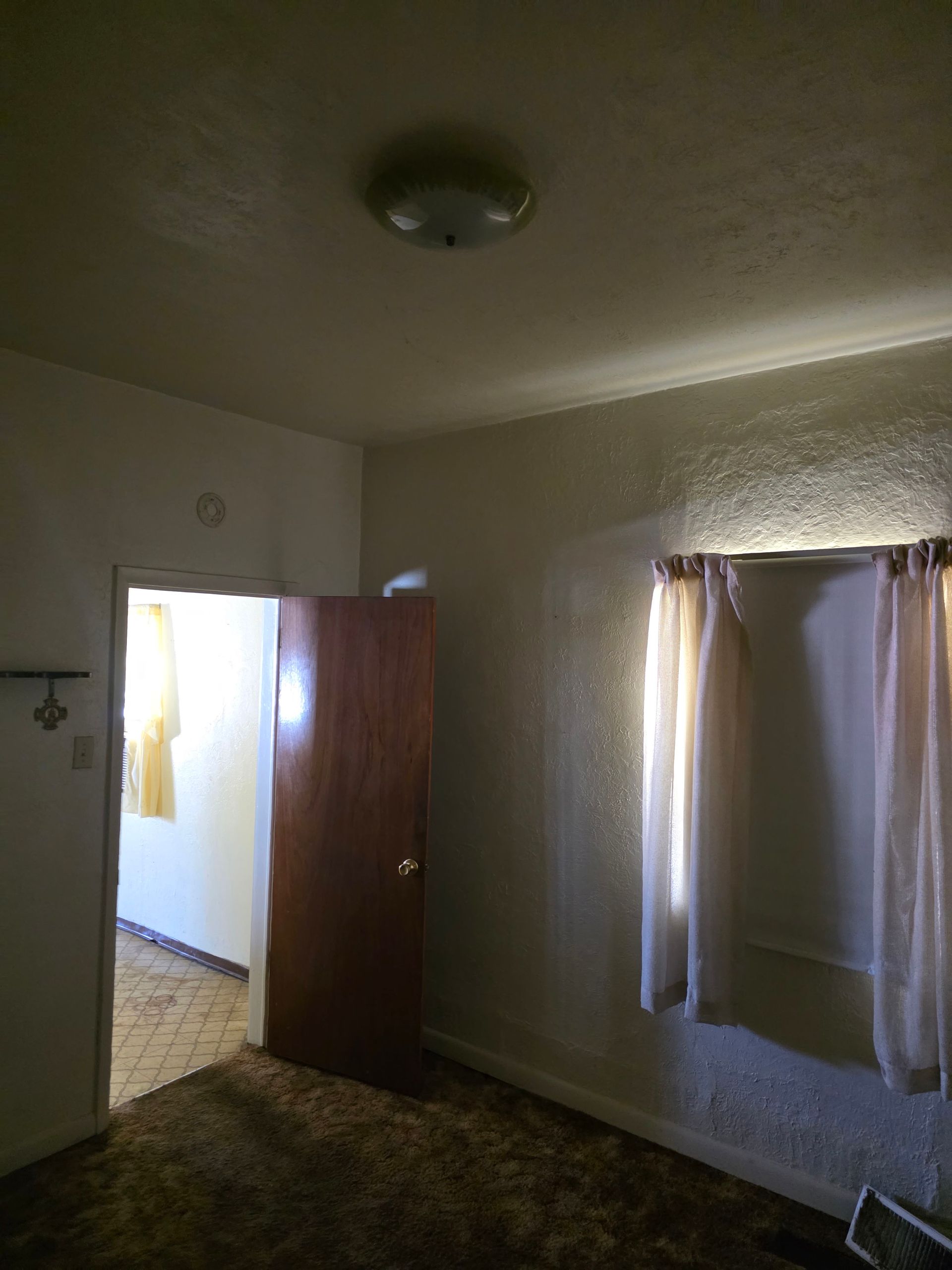 Small empty room with open door, curtained window, and ceiling light. Small empty room with open door, curtained window, and ceiling light.