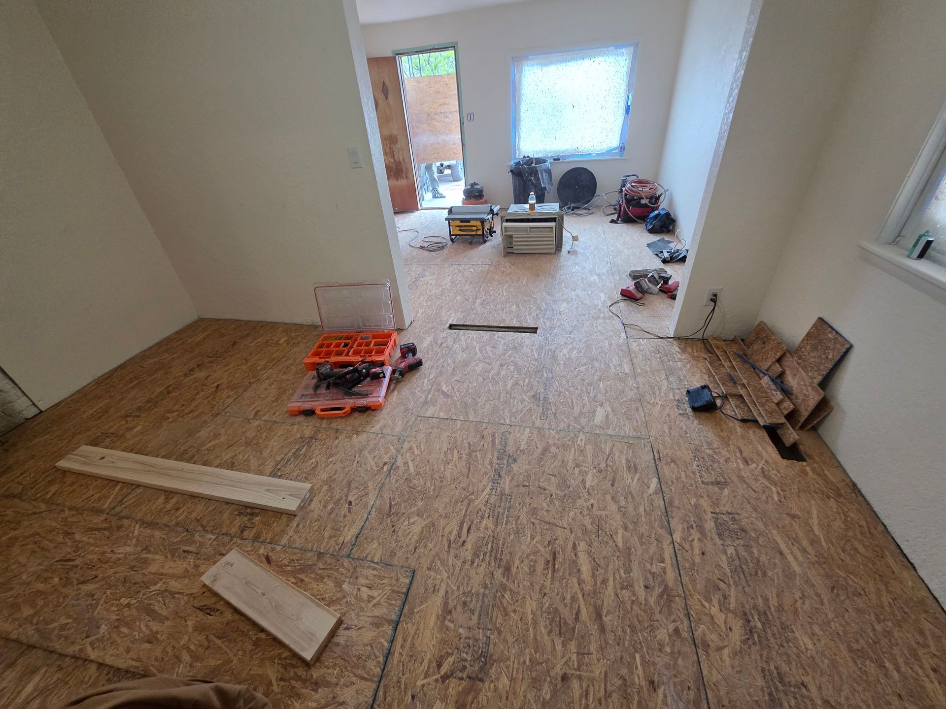 Sparse room under renovation with plywood subfloor, tools, and materials scattered near a bright window. Sparse room under renovation with plywood subfloor, tools, and materials scattered near a bright window.
