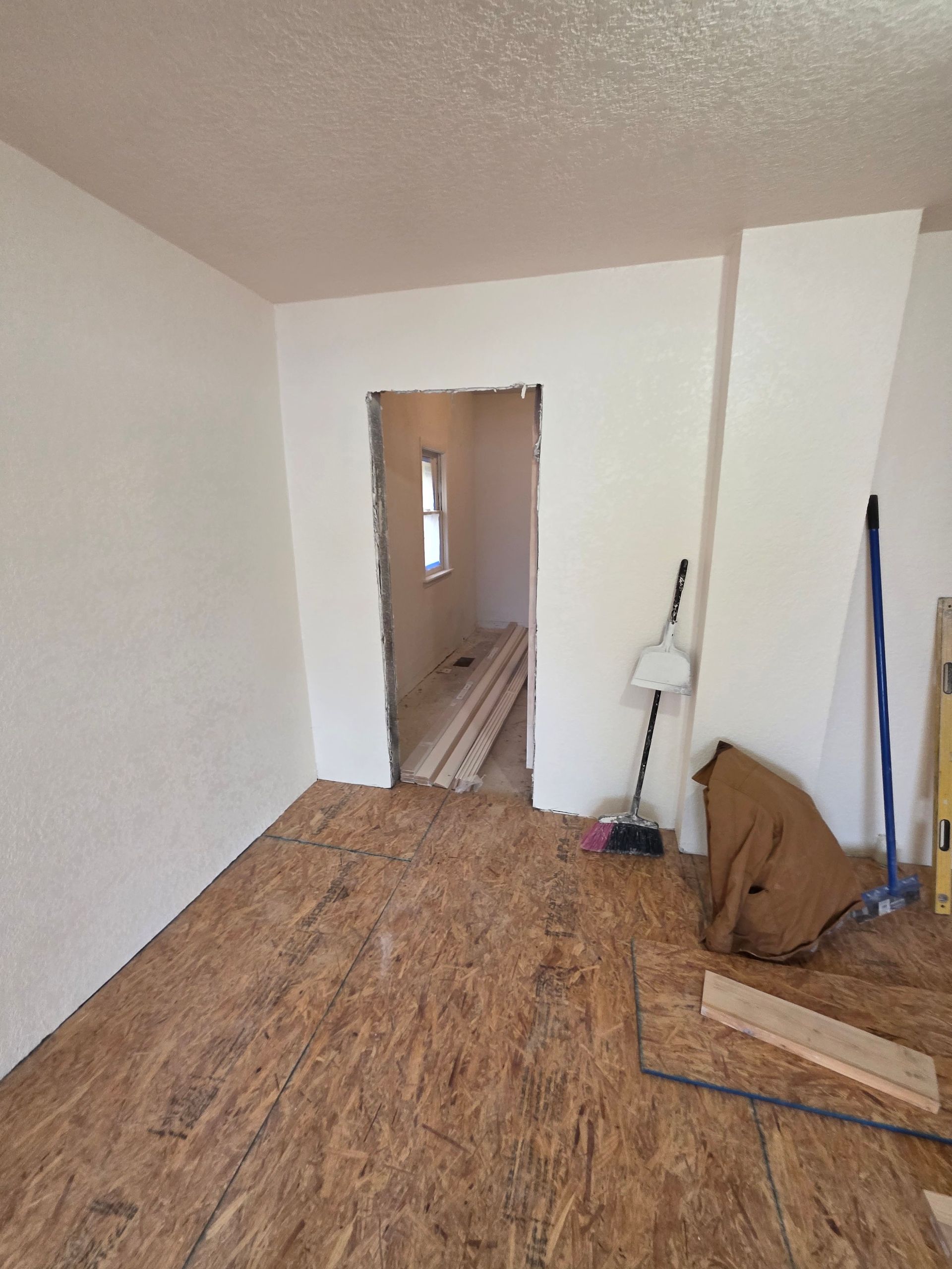 Bare room with plywood floor, white walls, open doorway, and tools leaning in the corner. Bare room with plywood floor, white walls, open doorway, and tools leaning in the corner.