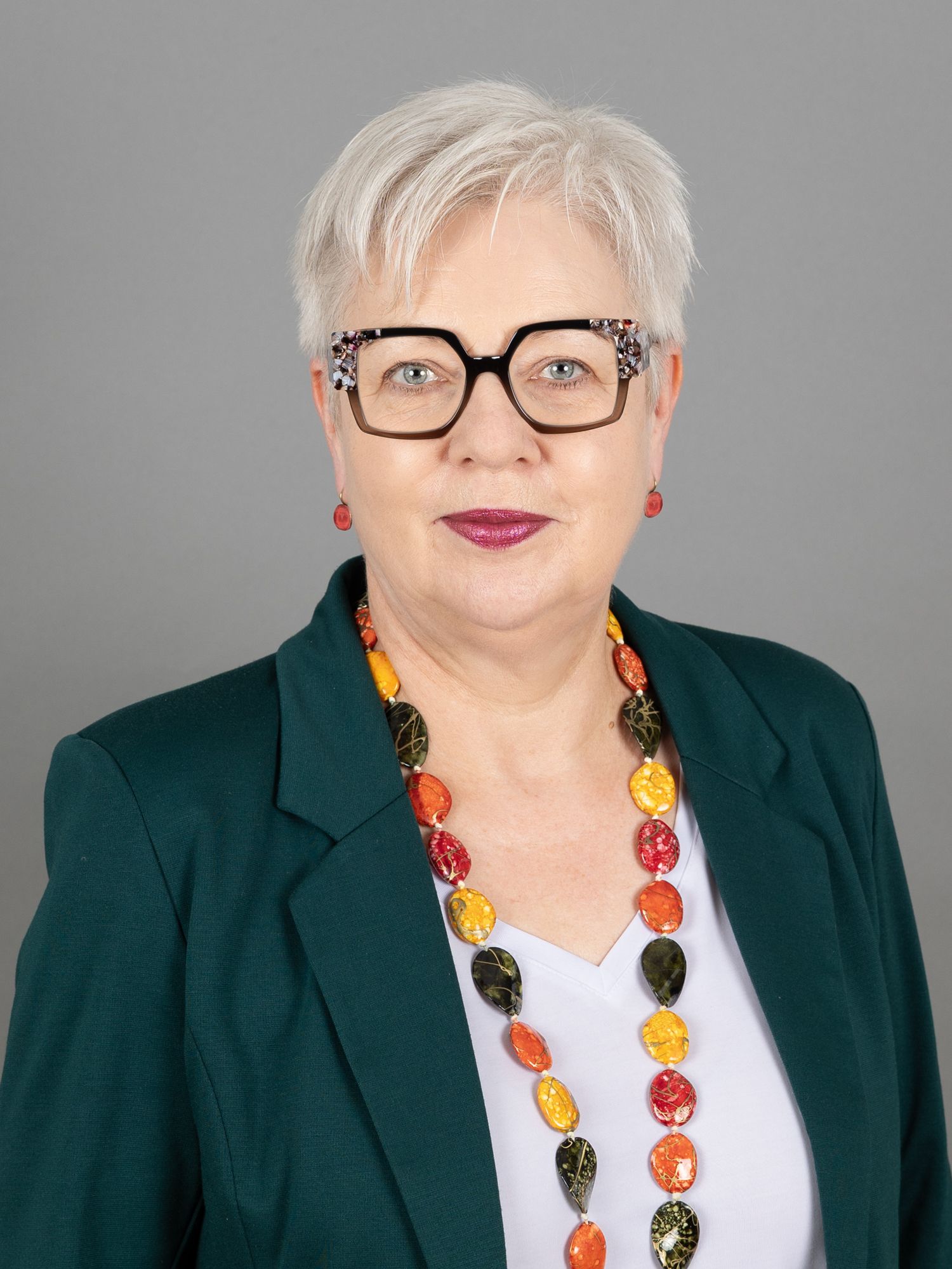 Frau mit Brille und bunter Halskette, die einen dunkelgrünen Blazer trägt.