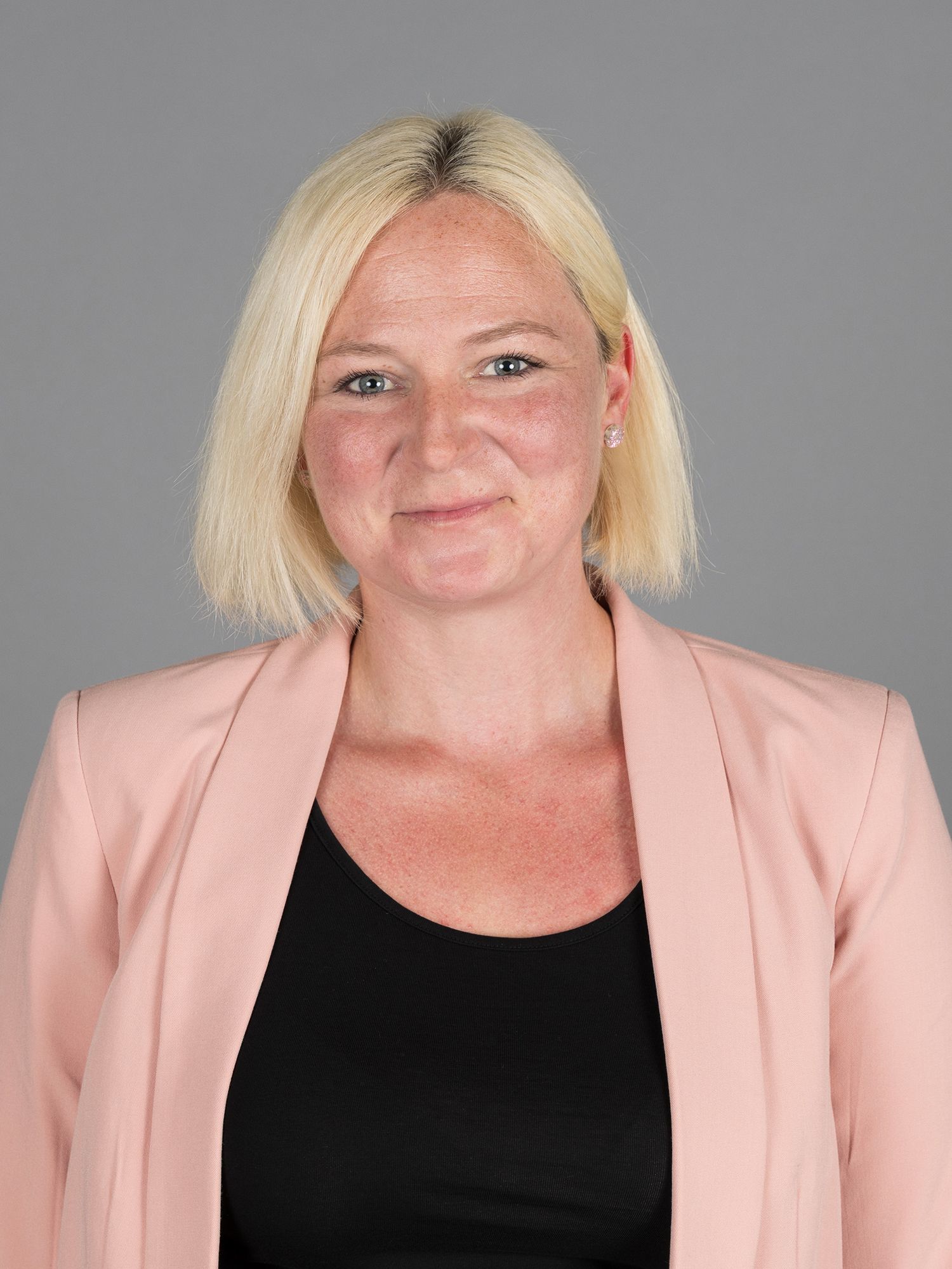 Frau in grüner Jacke und Brille lächelt, grauer Hintergrund.