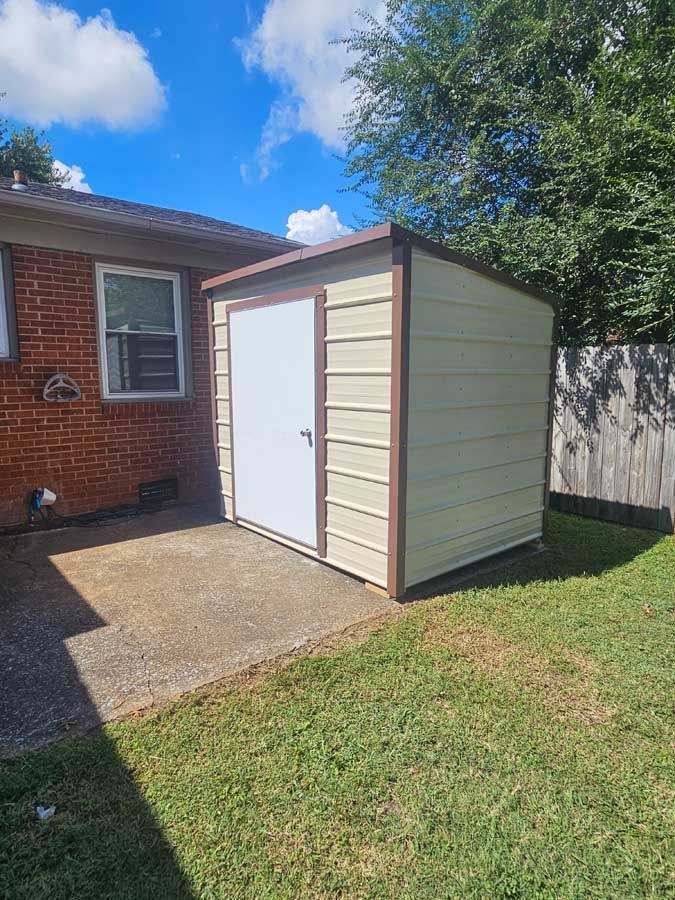 Tan shed with brown trim and white door, on concrete patio, near a brick building and green grass.