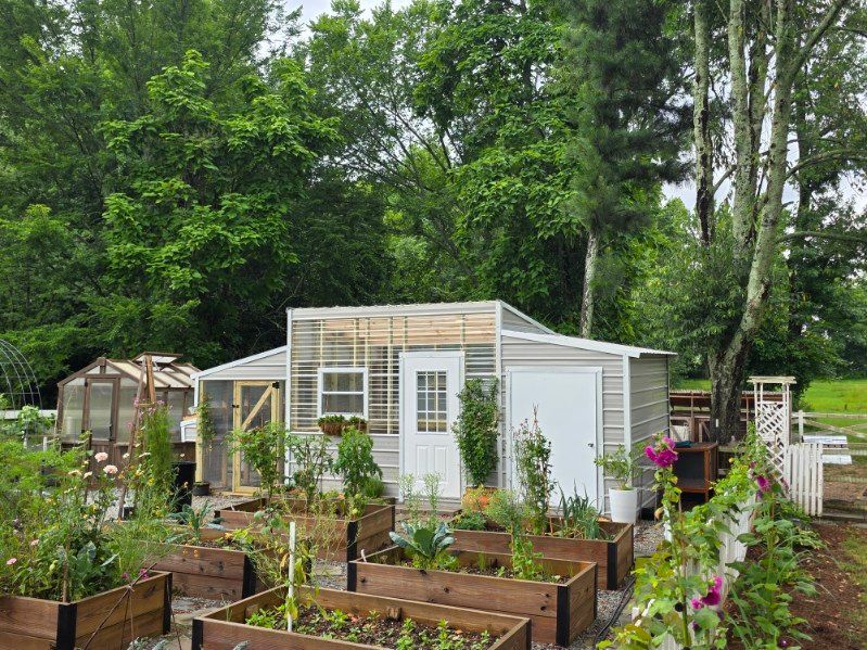 Raised garden beds and a white greenhouse in a lush green garden with trees in the background