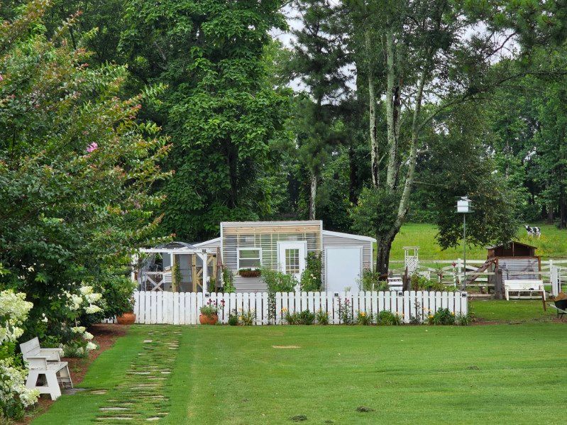 A white fenced garden with a small greenhouse, surrounded by trees and a green lawn.