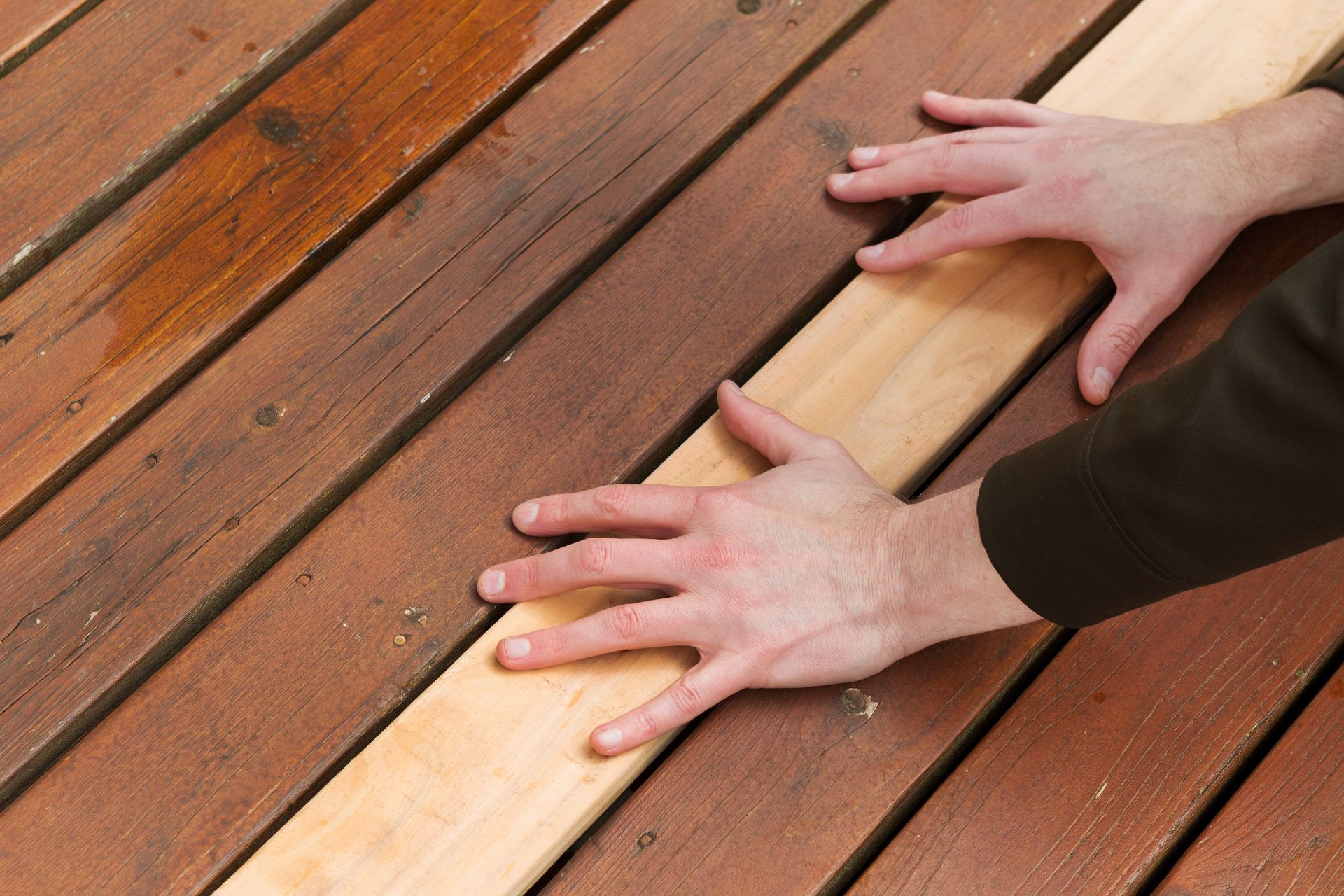 Hands arranging wooden planks on a deck, with one light-colored board among dark brown boards