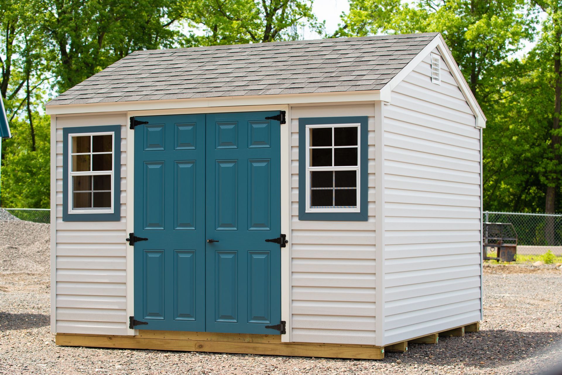 Small white shed with blue double doors and windows, set on gravel in a wooded yard
