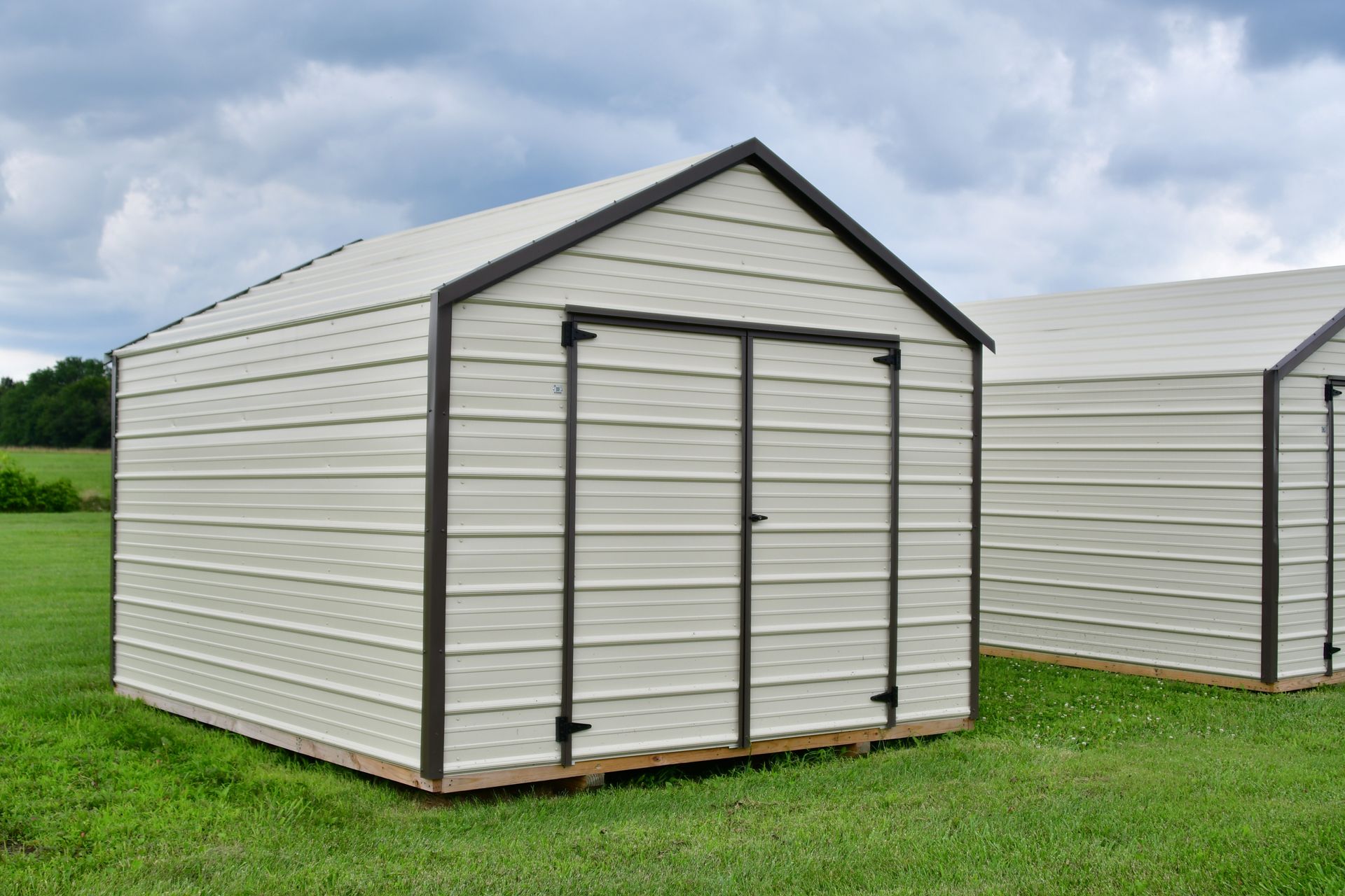 White metal storage shed with double doors on a grassy field, with another shed in the background