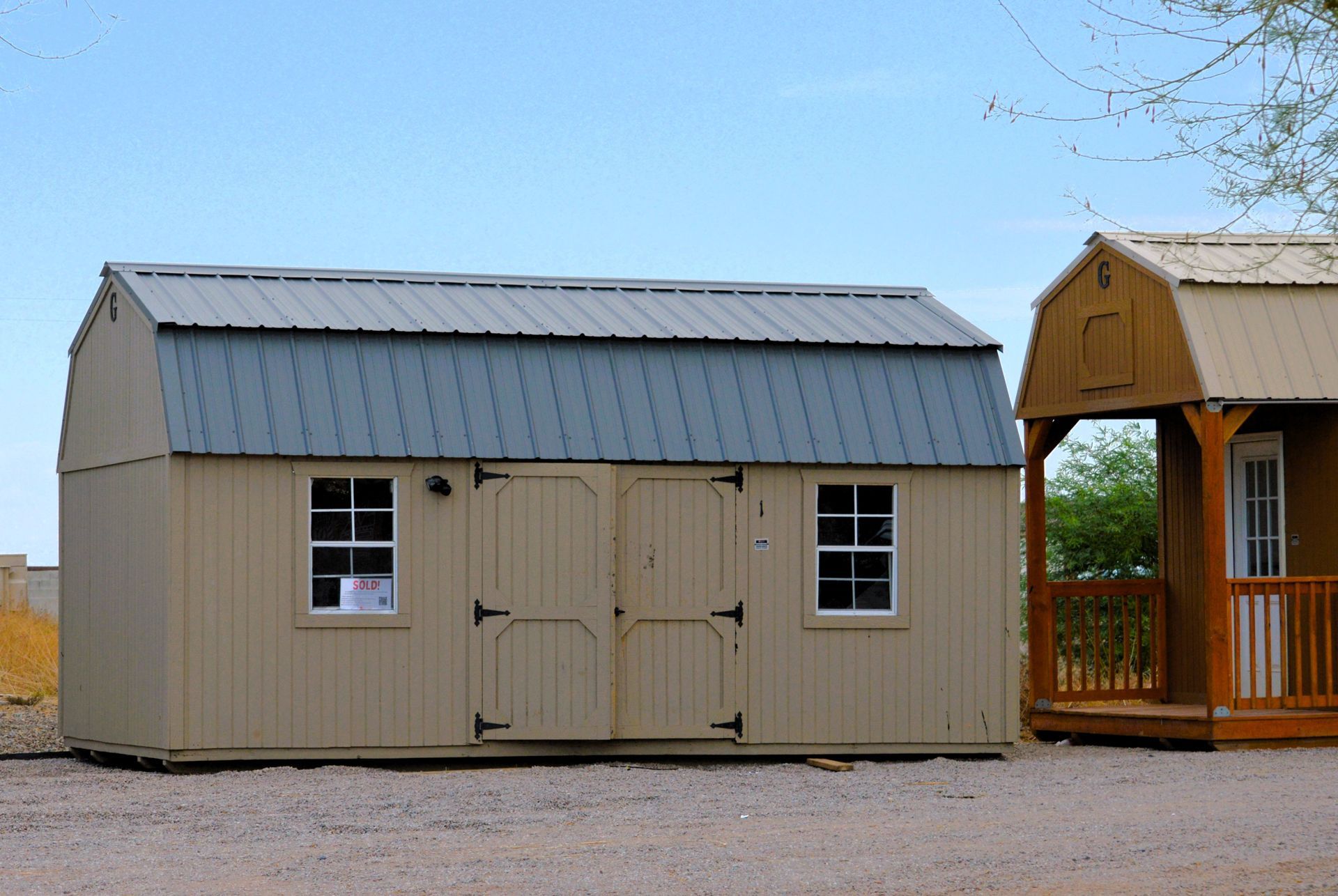 Tan shed beside a wooden gazebo on a gravel lot, with windows and double doors.