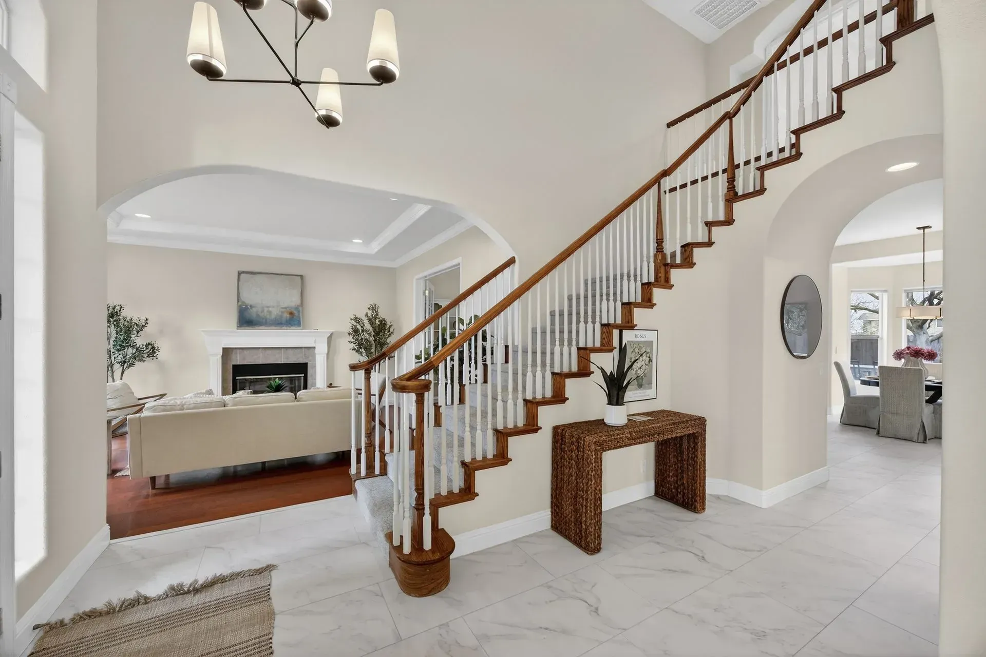 A bright, open foyer features a curved staircase with white spindles, a marble floor, and a view into a living room.