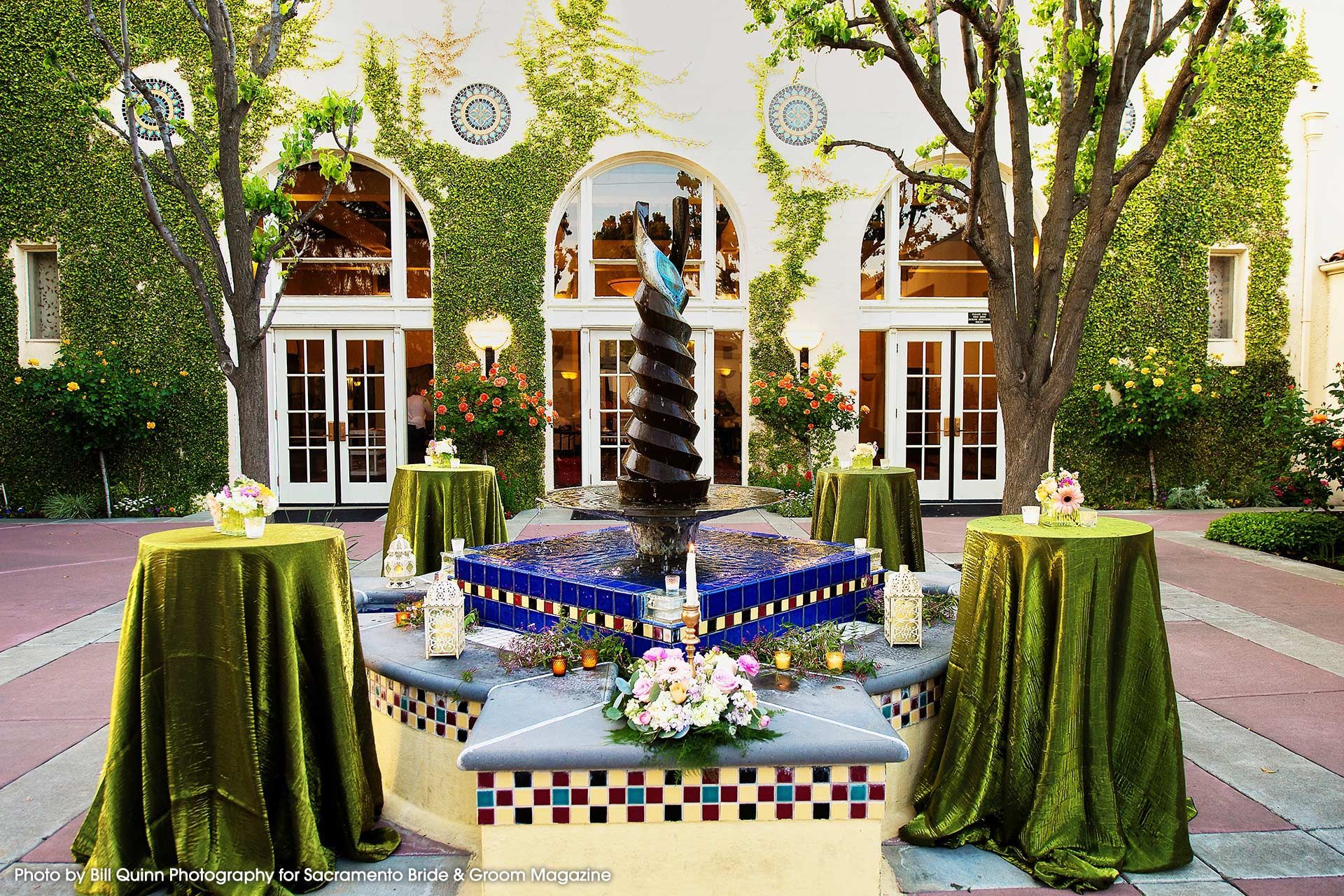 An outdoor patio features a central tiered fountain, draped cocktail tables with green cloths, and ivy-covered arches.