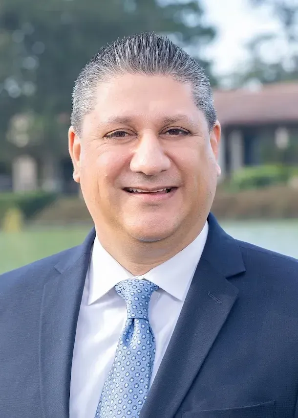 A man in a navy blue suit, white shirt, and light blue patterned tie posing for a portrait against an outdoor background.