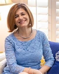 A smiling person with shoulder-length brown hair wearing a light blue lace dress and a pearl necklace, seated indoors.