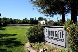 A sign for Stonegate Country Club next to a grassy field and trees on a sunny day.