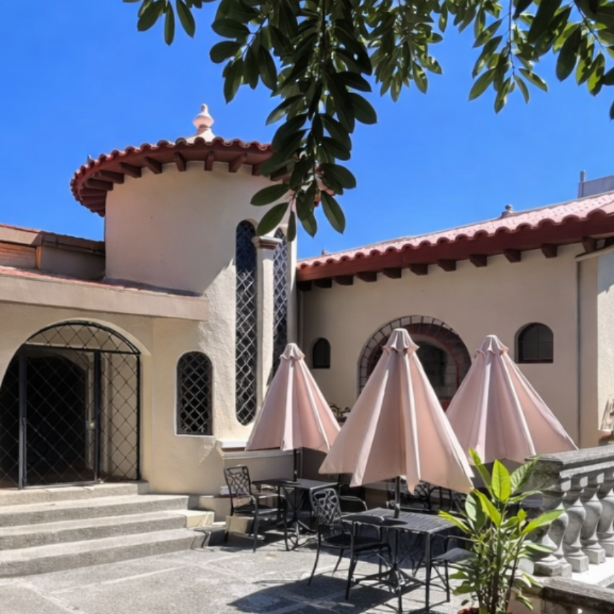 An outdoor patio with tables, chairs, and pink umbrellas next to a cream-colored building with arched windows and doors.