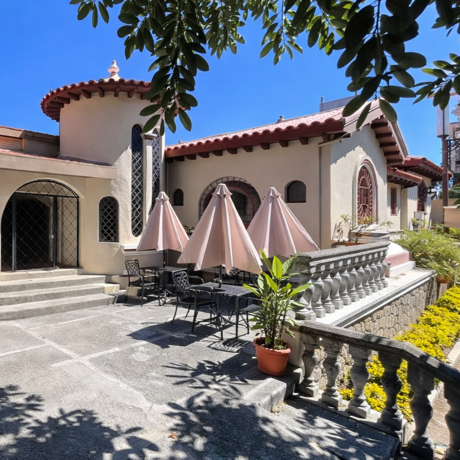 A sunny outdoor patio with tables, chairs, and beige umbrellas in front of a Spanish-style building with a stone terrace.