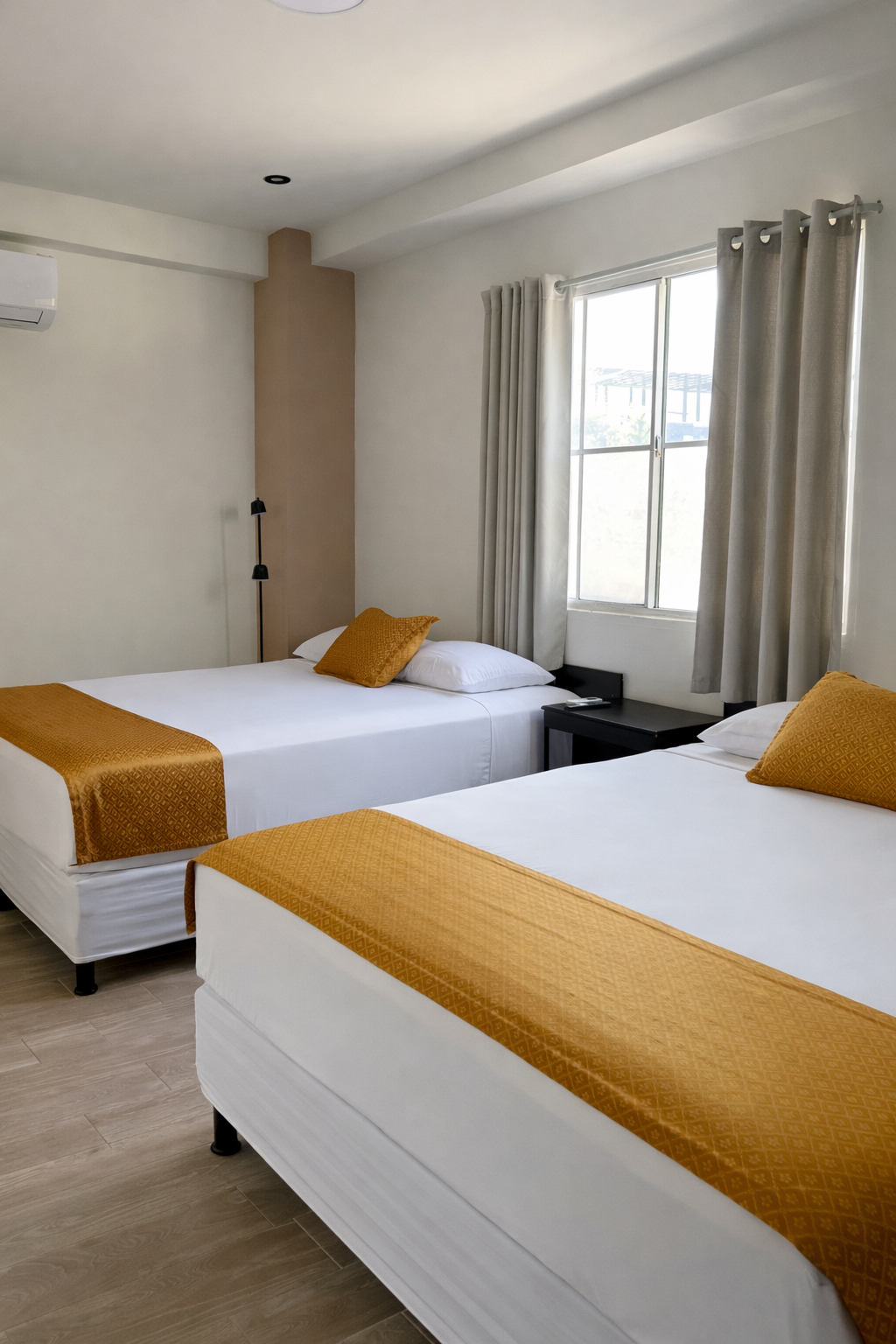 A bright hotel room with two white beds featuring mustard yellow runners and pillows, next to a window with gray curtains.