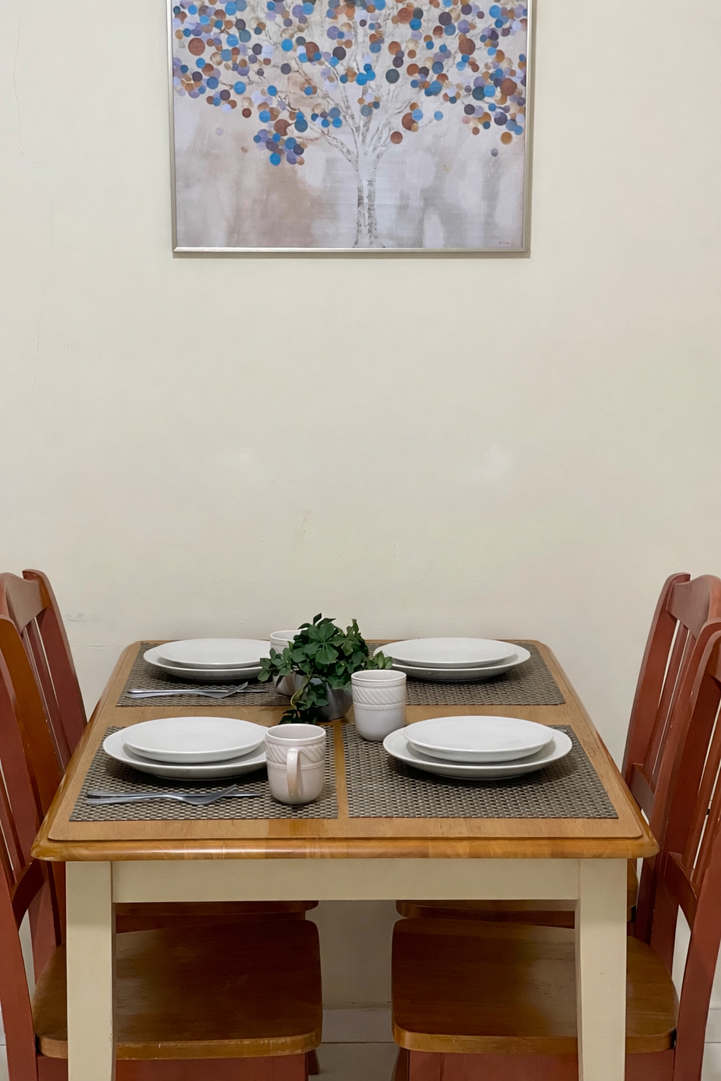 A four-person wooden dining table with place settings, a small plant, and framed artwork on the wall above.