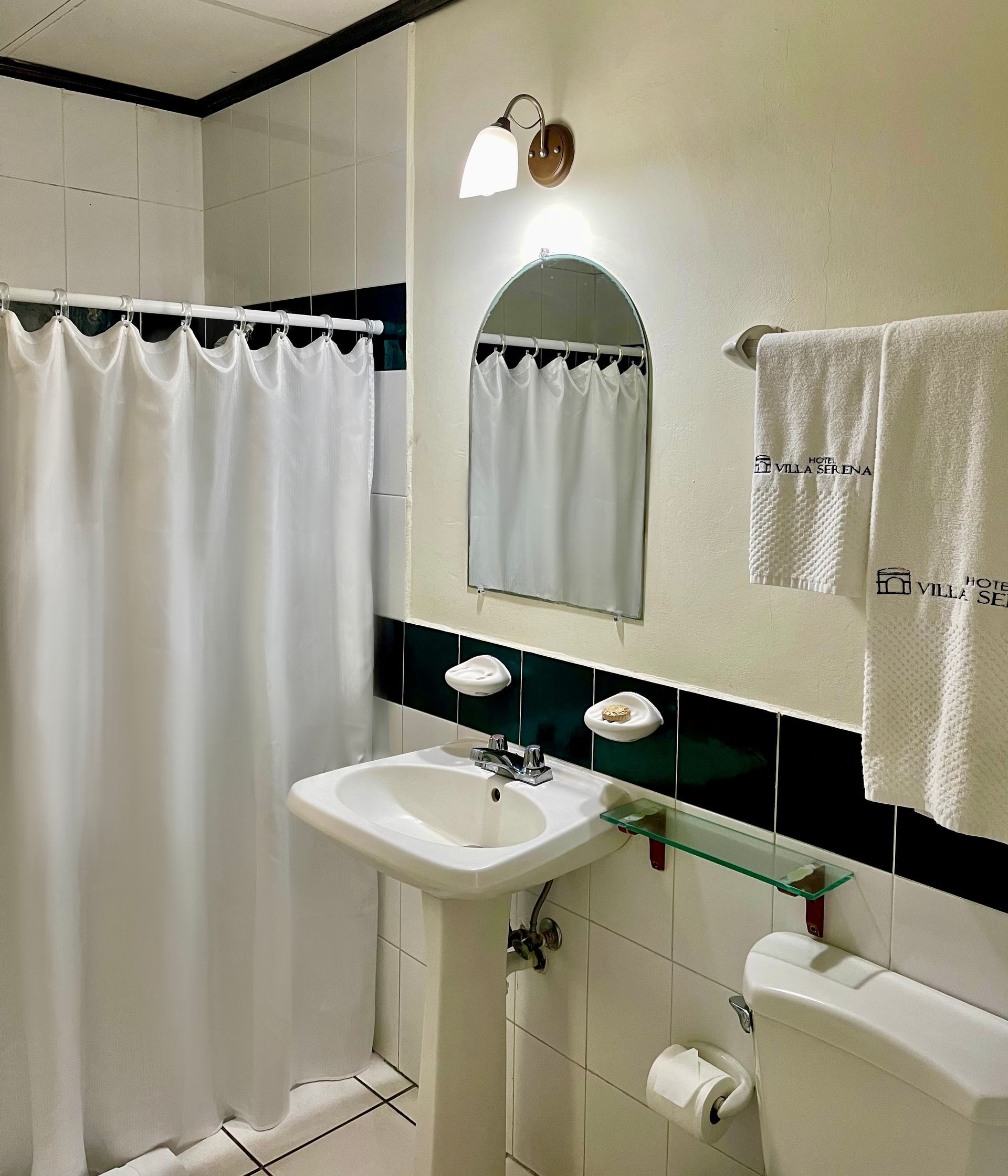 A bathroom with a pedestal sink, a mirror with a light fixture above it, a shower with a white curtain, and a toilet.