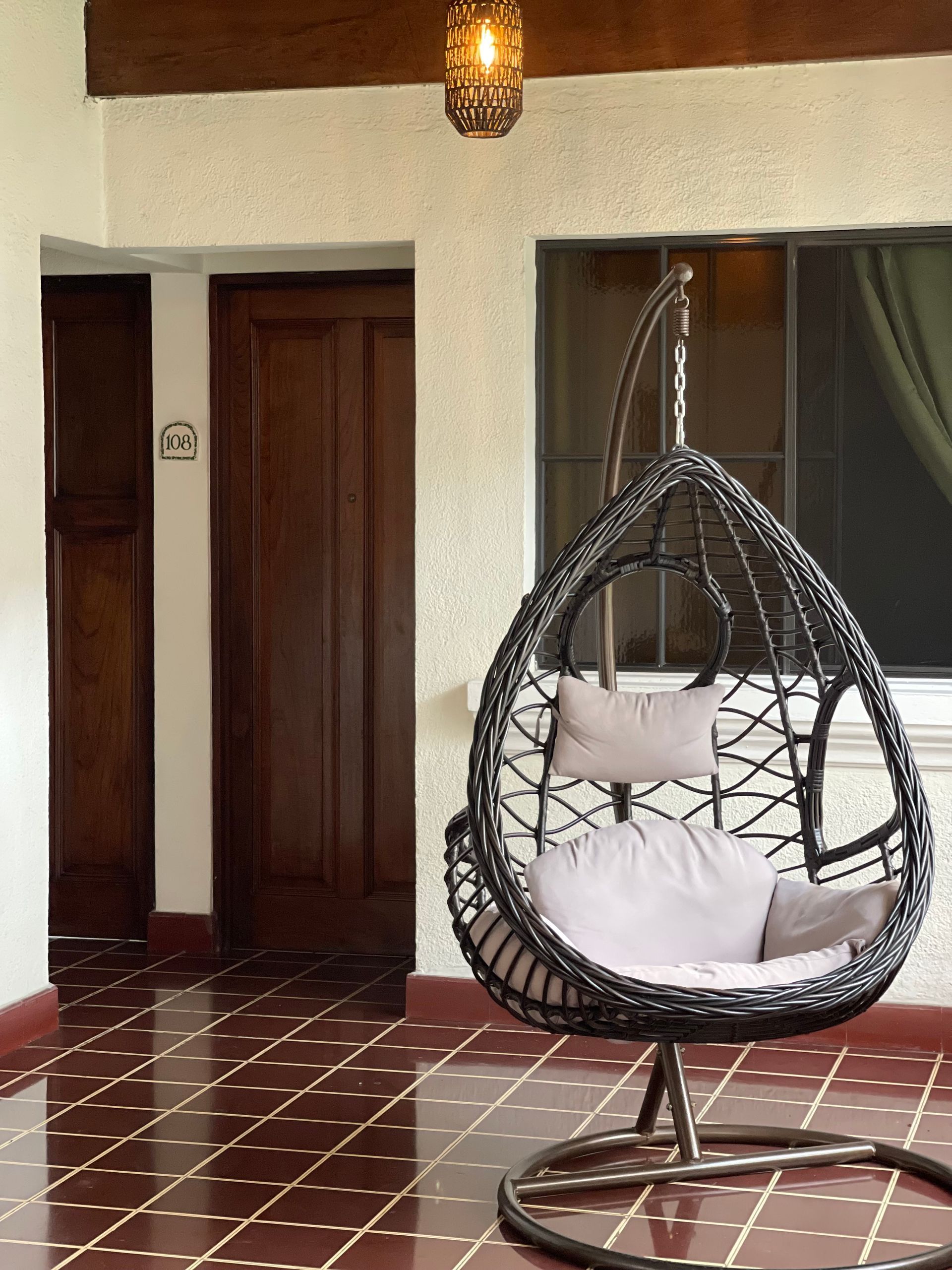 A hanging wicker chair with light-colored cushions stands on a red-tiled floor in a room with wood doors and a pendant light.