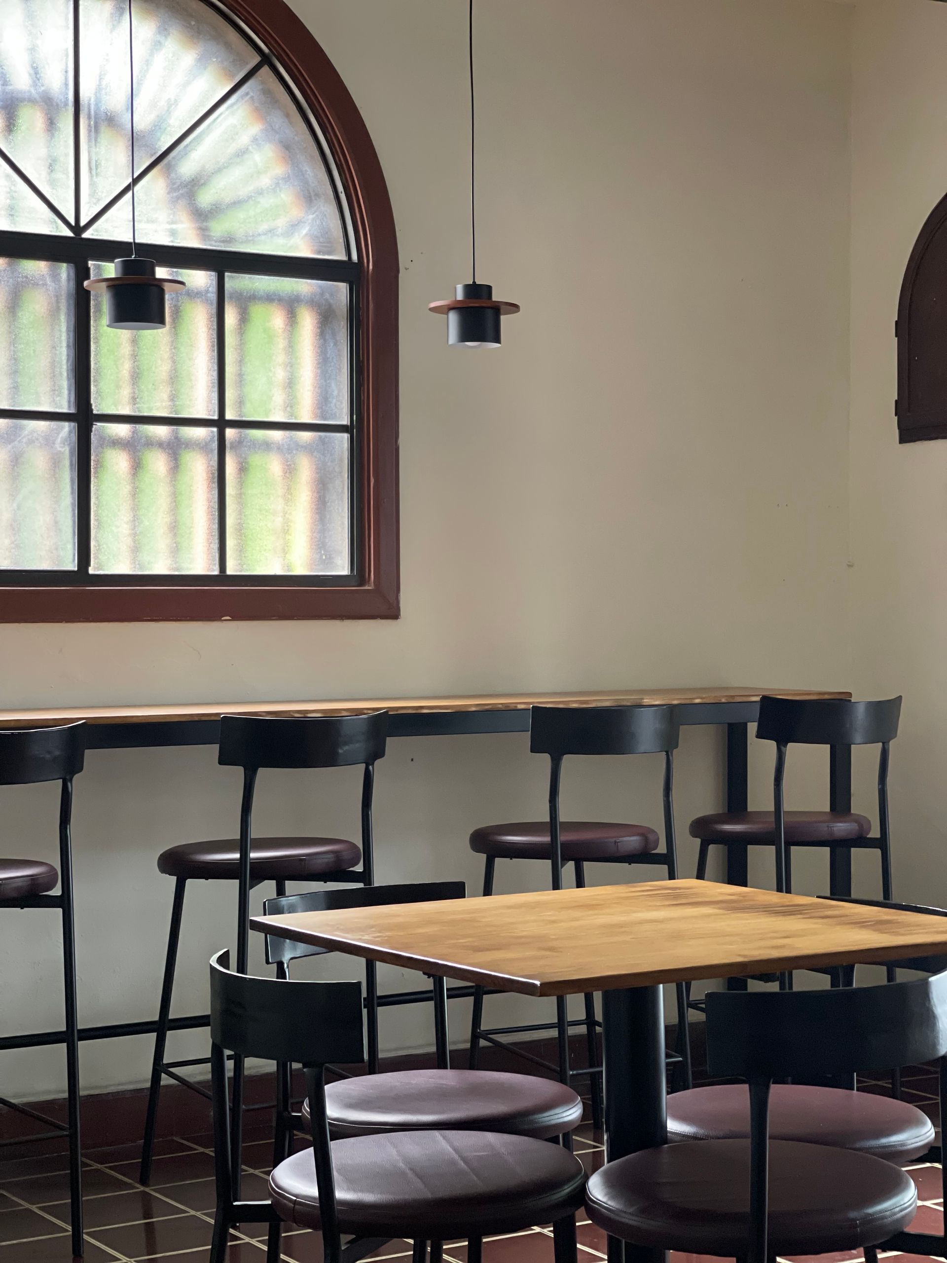 Empty chairs are positioned around a wooden table and counter in a cafe with a large, arched window.