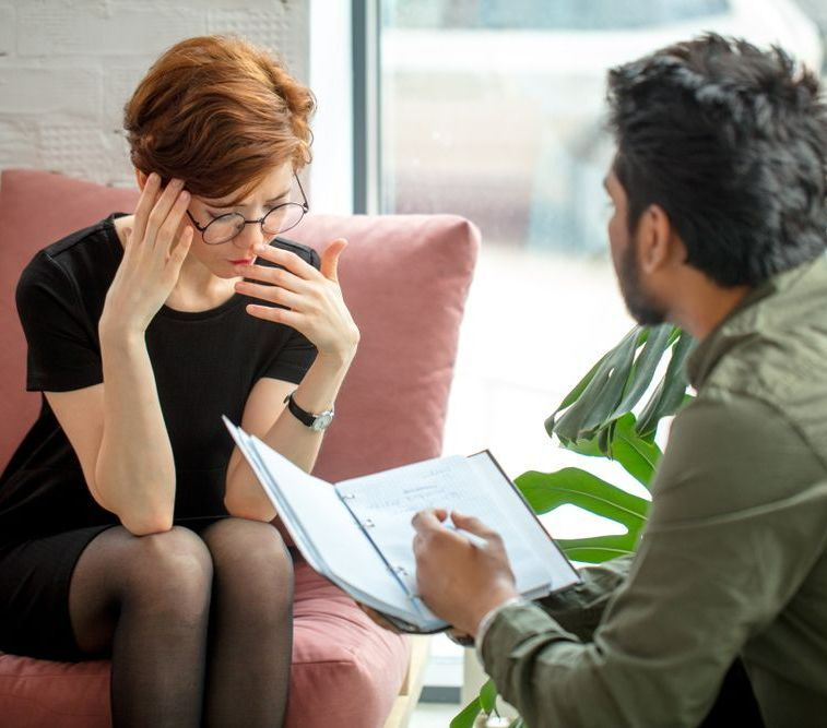 A Woman Is Sitting In A Chair Talking To A Man Who Is Holding A Clipboard — The Therapists Cairns In Cairns City, QLD