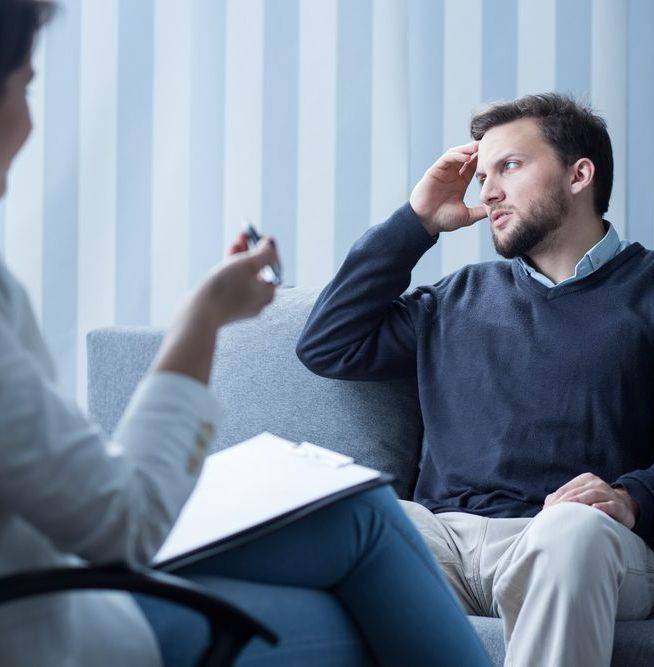 A Man Is Sitting On A Couch Talking To A Woman Who Is Holding A Clipboard — The Therapists Cairns In Cairns City, QLD