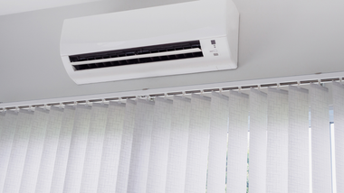 A white wall-mounted air conditioner positioned above white vertical blinds on a ceiling track.