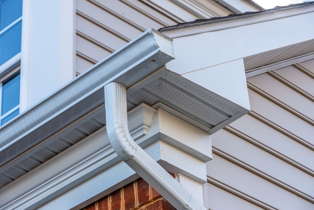 White vinyl siding and soffit with a white metal gutter and a downspout attached to the corner of a house.