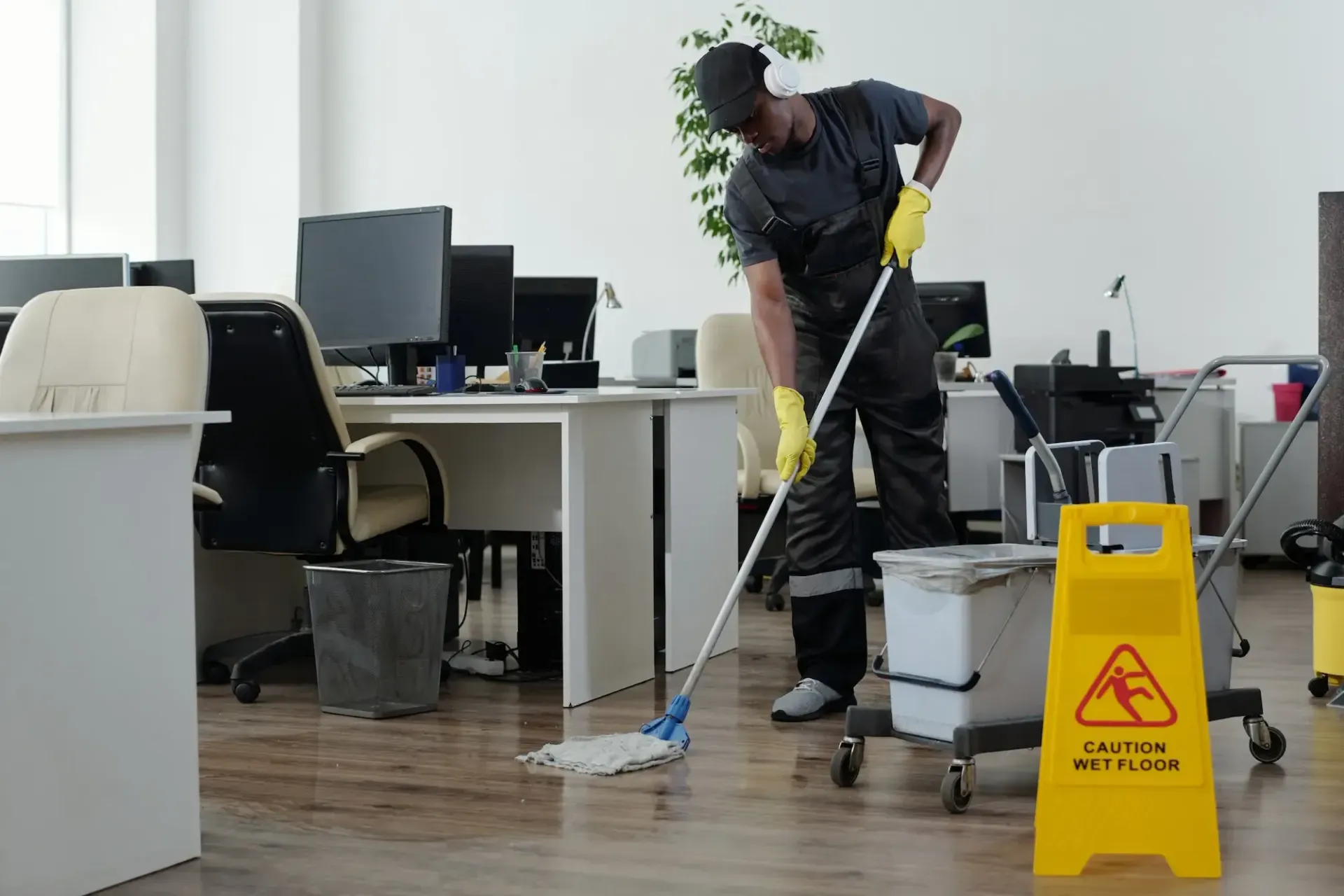 Une personne nettoie le sol d'un bureau, un panneau jaune « Attention sol mouillé » se trouvant à proximité.