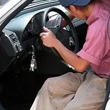 A person in a pink shirt cleans the interior dashboard of a car with a cloth while holding the steering wheel.