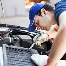 A mechanic wearing a blue cap and work gloves uses a wrench to repair a car engine in a garage.