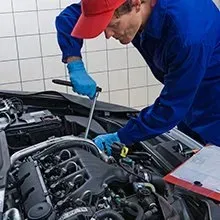 A mechanic in a blue uniform and red cap uses a wrench to work on a car engine while referring to a clipboard.