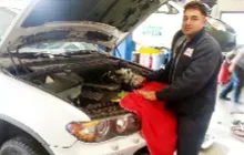A mechanic wearing a dark jacket leans over the open hood of a white vehicle, holding a red cloth in a repair shop.