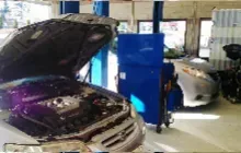 A car with an open hood revealing a purple engine block inside a garage, with another vehicle parked in the background.