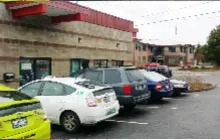 A parking lot in front of a tan commercial building with a red roof, featuring several cars parked in a row.