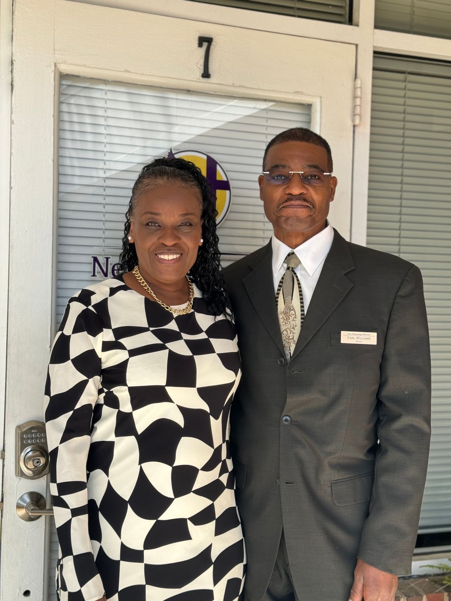 Two people standing at a doorway marked “7,” wearing formal black-and-white outfits and smiling.