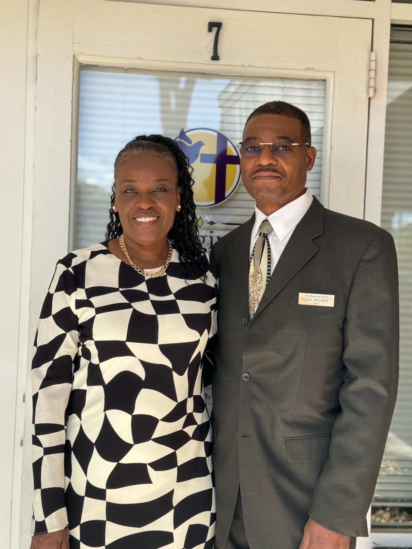 Two people stand smiling in front of a doorway marked “7,” dressed in formal attire.