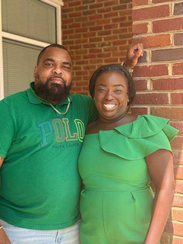 Two people in green outfits smiling on a brick porch, standing close together.