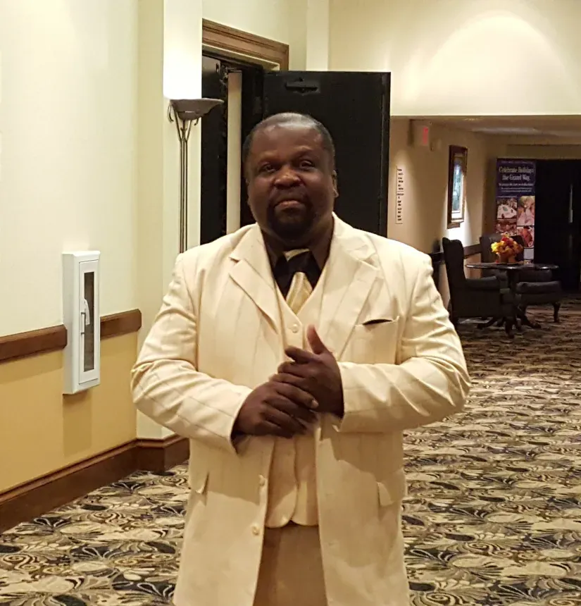 Man in a cream suit standing in a hotel hallway with hands clasped, looking at the camera.