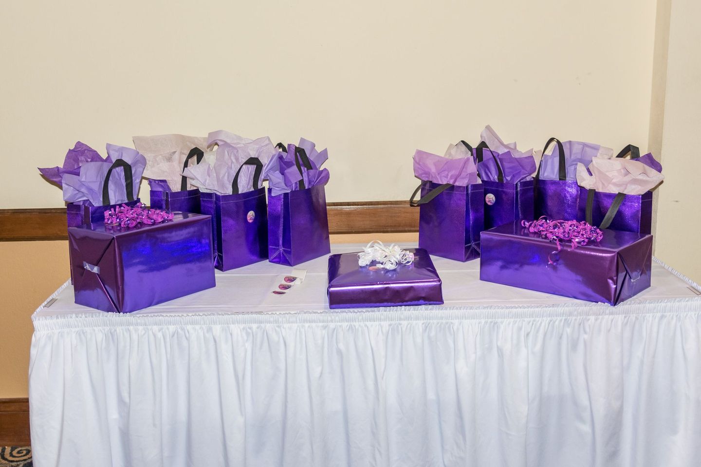 Purple gift bags and a small wrapped box arranged on a table with a white tablecloth