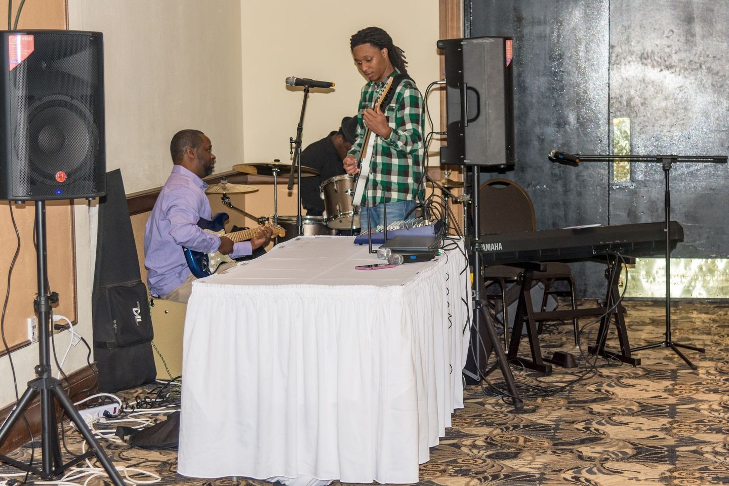 Musicians performing indoors beside speakers, a keyboard, and a microphone stand with a table in front.
