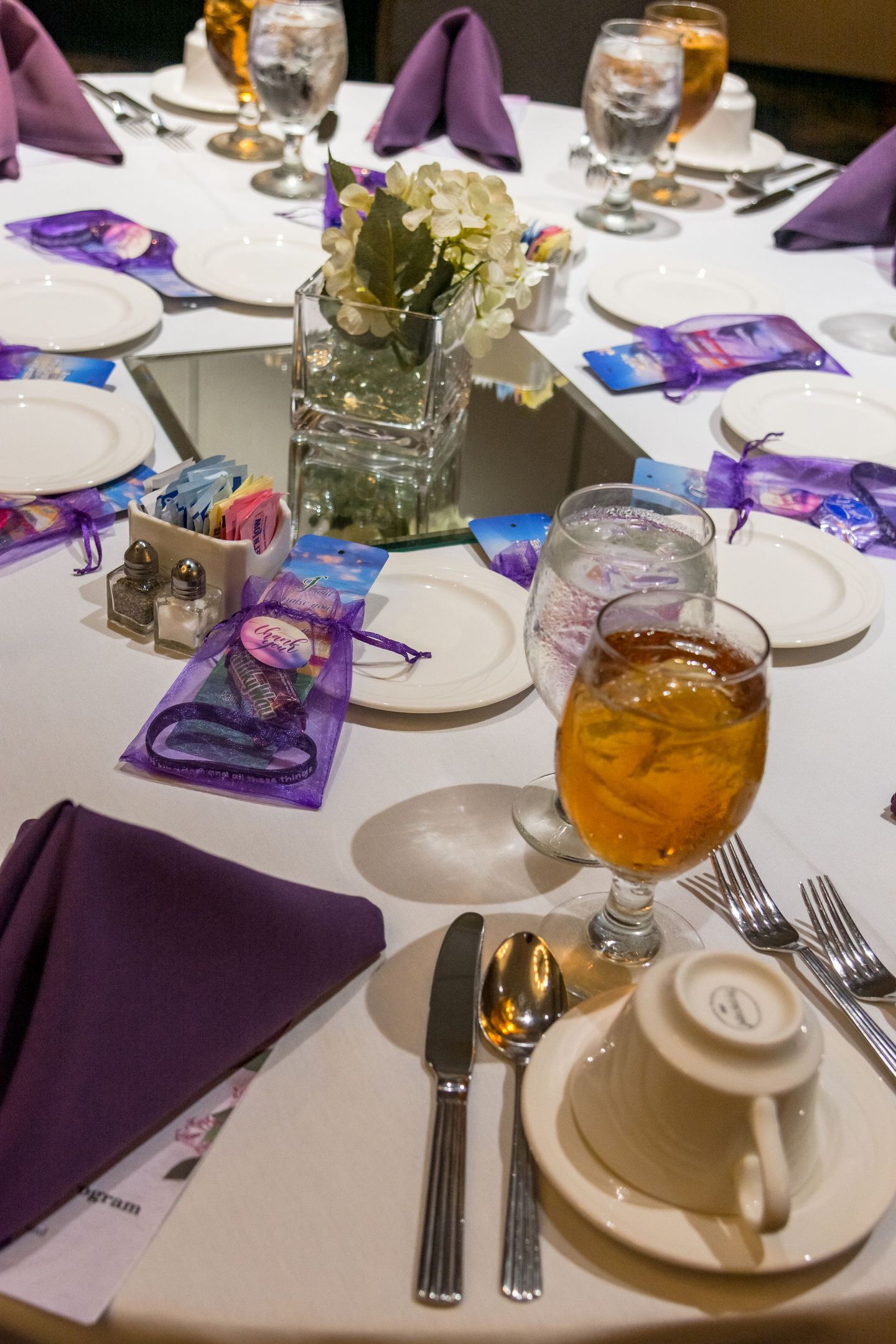 Banquet table set with purple napkins, glassware, plates, and iced drinks, centered by a floral vase.