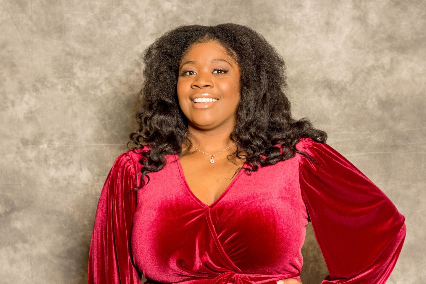 Smiling person in a red velvet dress against a textured gray backdrop