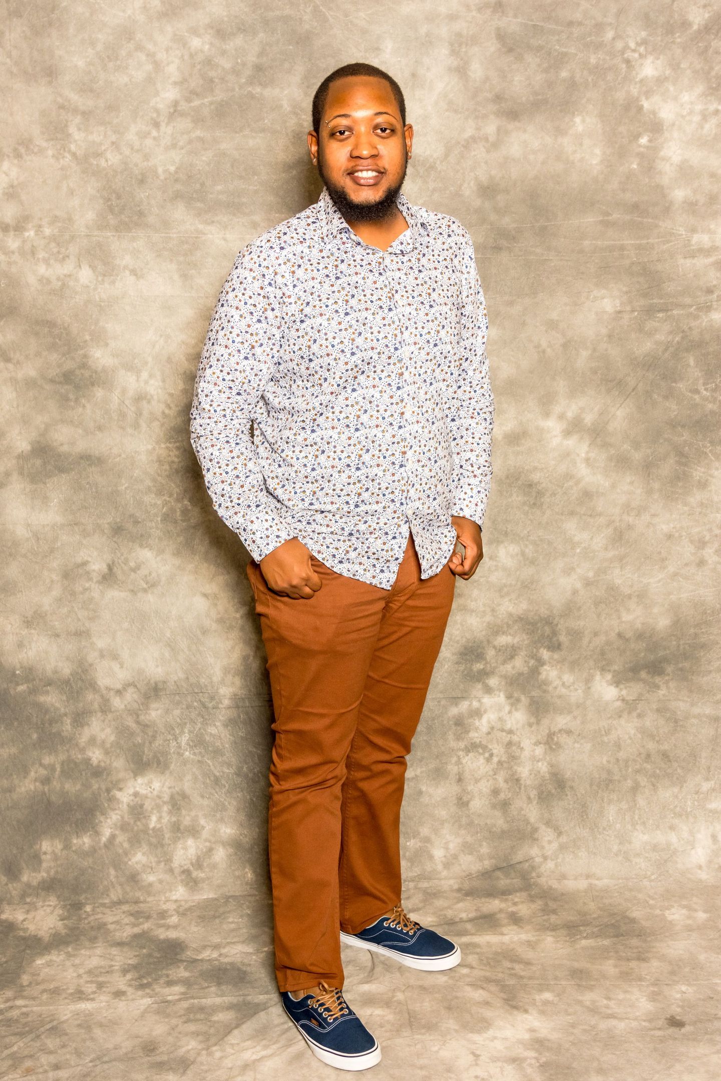 Person standing against a gray backdrop, wearing a white patterned shirt, brown pants, and blue shoes.